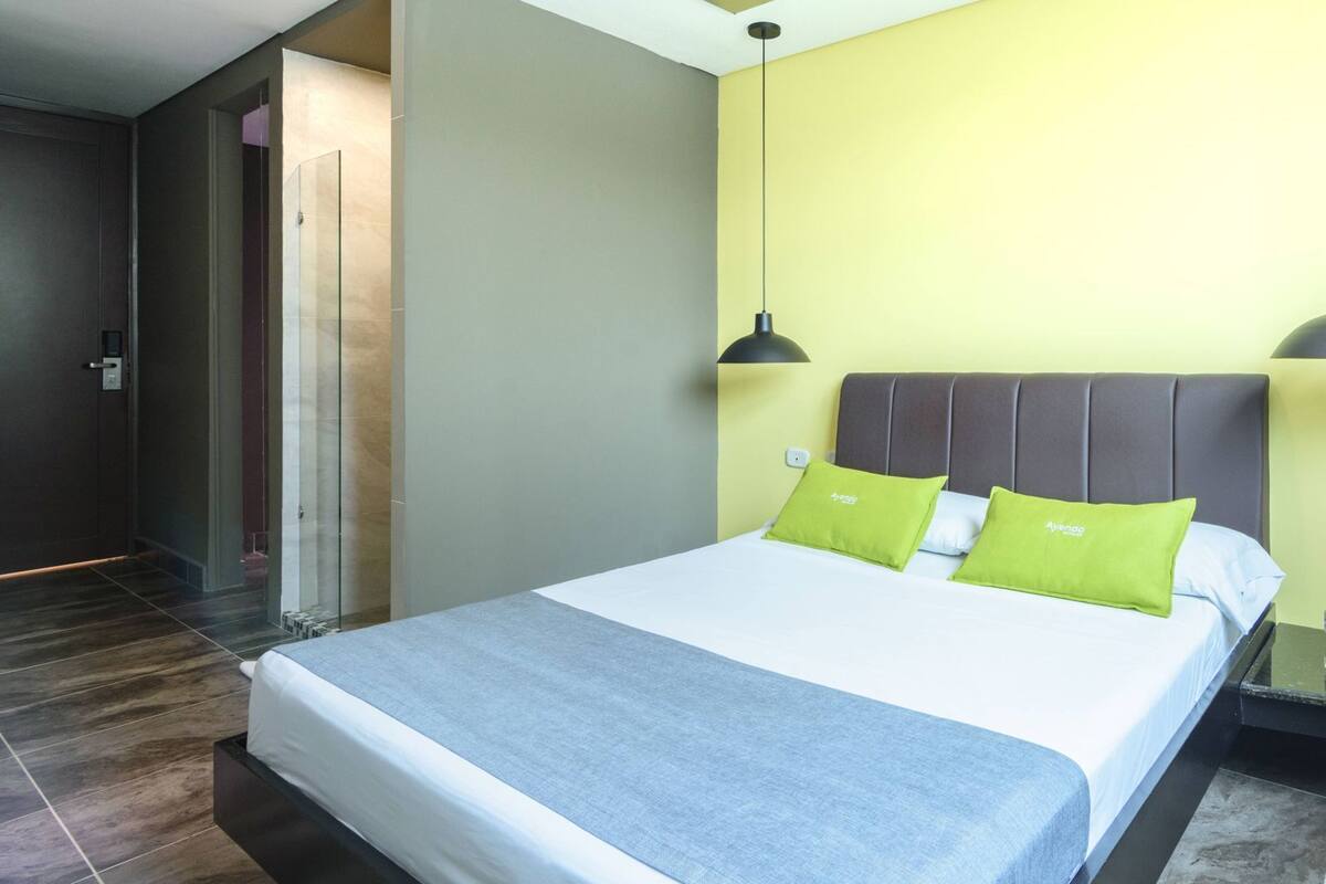 A modern hotel room with a bed, a nightstand, a lamp, and a door leading to a bathroom.