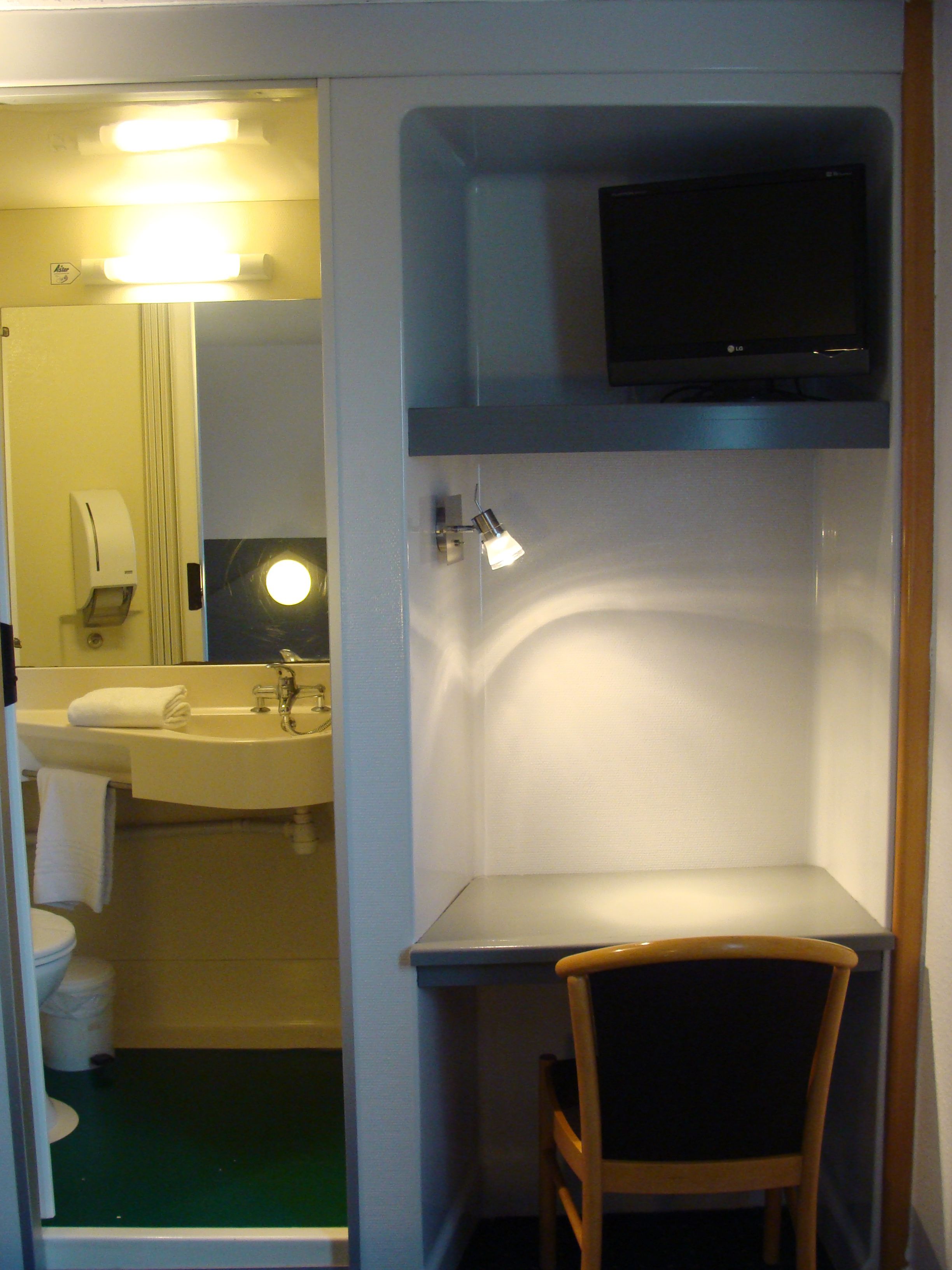 Une chambre d’hôtel compacte avec une salle de bain comprenant un lavabo, un miroir et des toilettes. On y trouve également une petite chaise en bois et un téléviseur à écran plat fixé au mur.