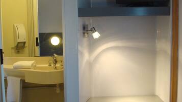 Une chambre d’hôtel compacte avec une salle de bain comprenant un lavabo, un miroir et des toilettes. On y trouve également une petite chaise en bois et un téléviseur à écran plat fixé au mur.