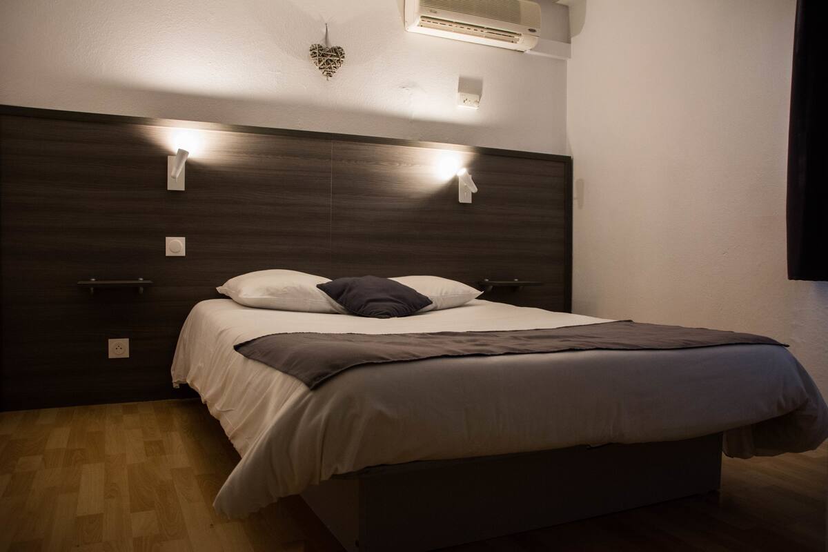 Une chambre à coucher moderne avec une tête de lit en bois foncé, un couvre-lit blanc et deux lampes de lecture fixées au mur.