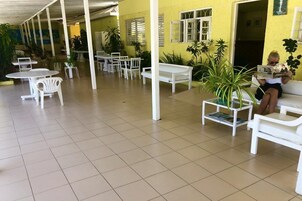 Un espacio de estar al aire libre techado, con mesas y sillas blancas, rodeado de macetas con plantas.