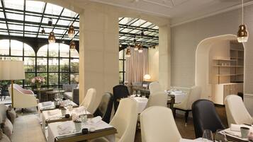 Un restaurant avec de hauts plafonds, de grandes fenêtres et des lustres modernes. Les tables sont dressées avec des nappes blanches et de beaux couverts.