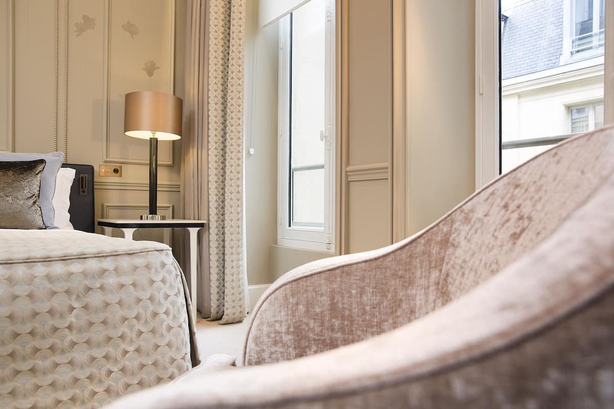 Habitación de hotel con una cama, una silla, una lámpara, una mesita y una ventana con vistas a edificios.