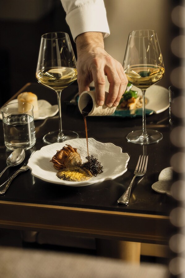Una mesa de comedor con un plato de comida, una copa de vino blanco y una mano sirviendo salsa.