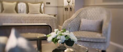 A well-lit hotel room with a bed, a sofa, a glass coffee table with a vase of flowers, and a wall-mounted light fixture.