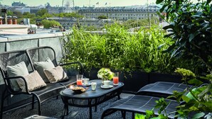 Terraza en la azotea con mobiliario exterior, una mesa con bebidas y aperitivos, y vistas a un paisaje urbano que incluye una noria.