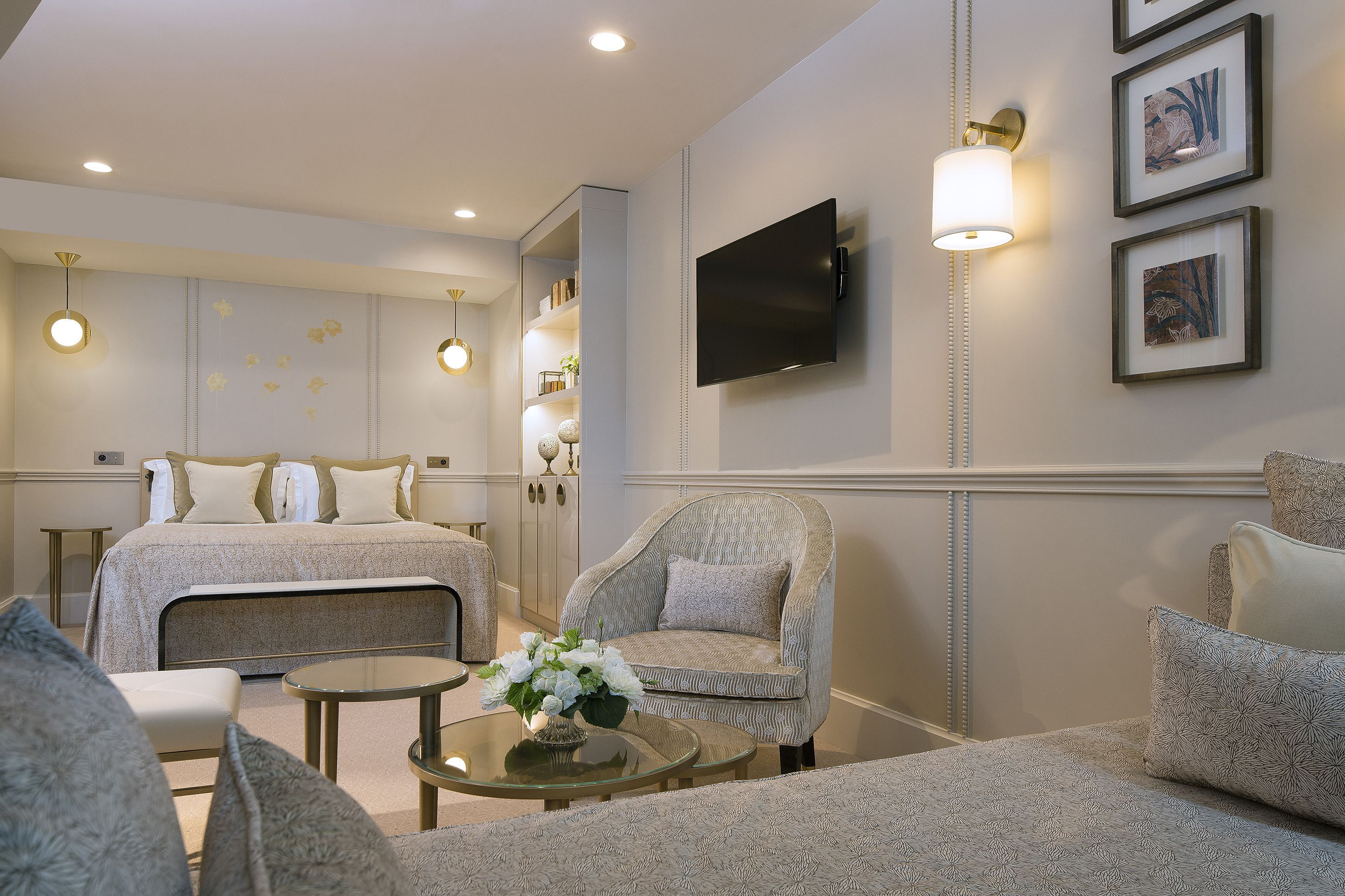 A hotel room with a bed, a sofa, a small table with flowers, a TV mounted on the wall, and framed artwork.