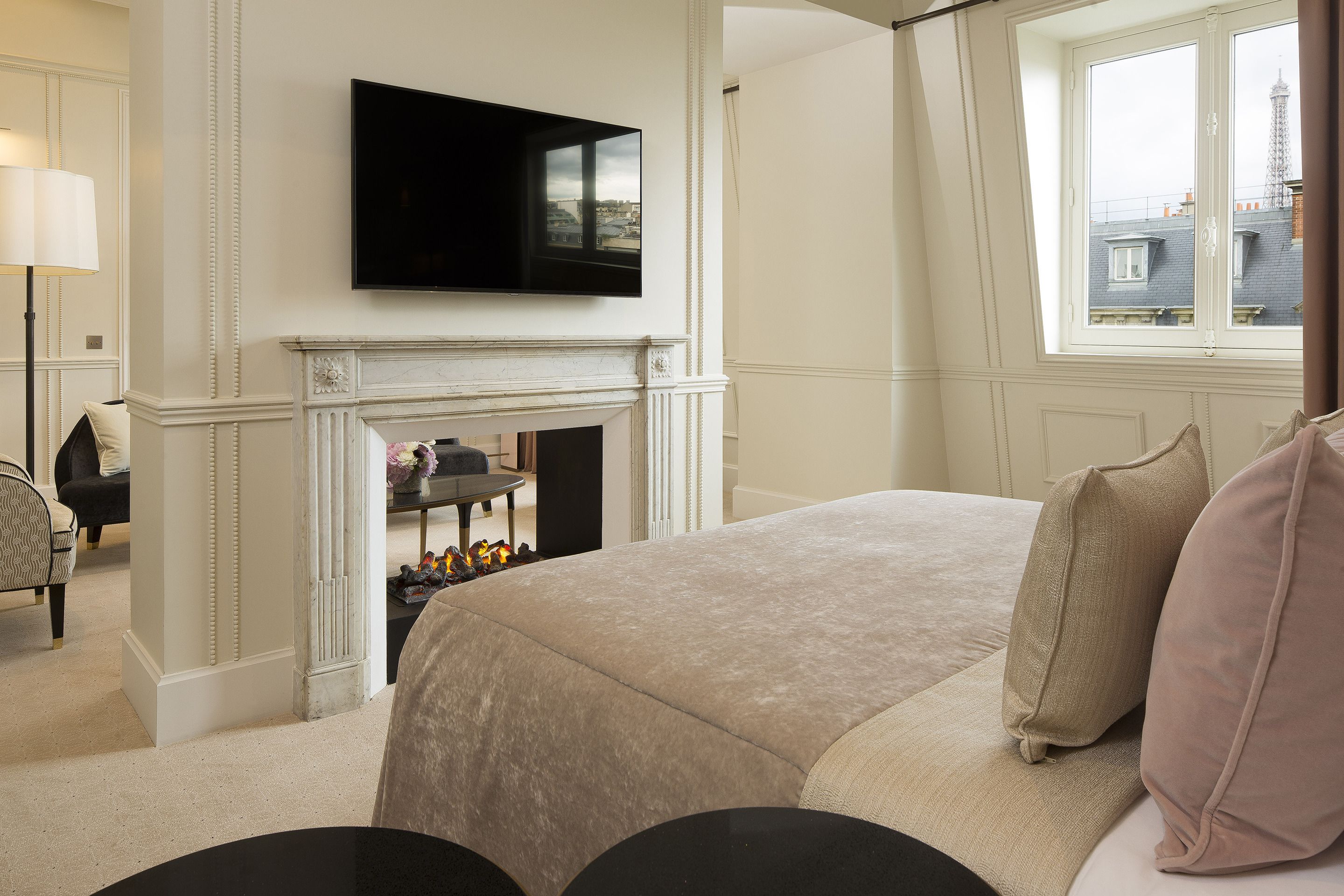 Un dormitorio con una cama grande, un televisor de pantalla plana sobre una chimenea y vistas de edificios a través de una ventana.