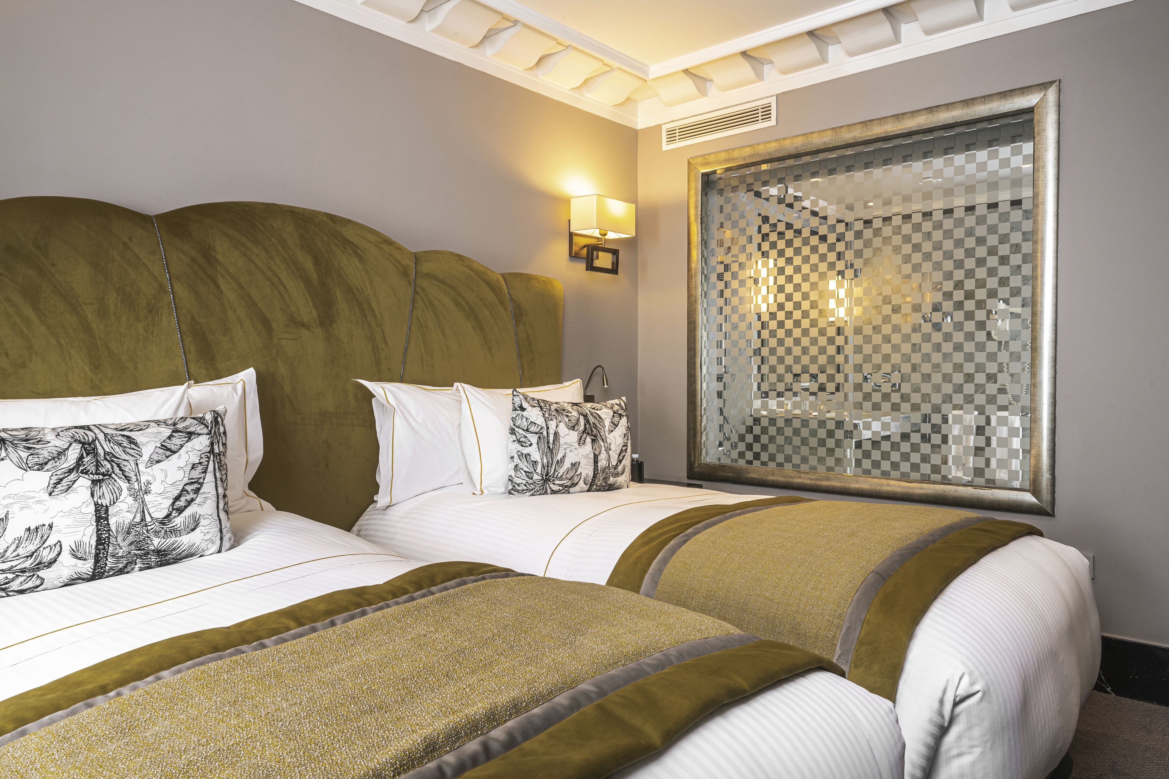 A hotel room with two beds, each with white bedding and decorative pillows, a large mirror with a checkered pattern, and a wall-mounted light fixture.