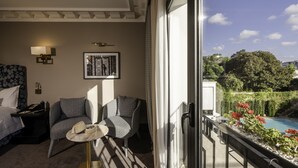 Habitación de hotel con cama, dos sillones, una mesita, un cuadro enmarcado, una mesita de noche y vistas a una piscina y vegetación al exterior.