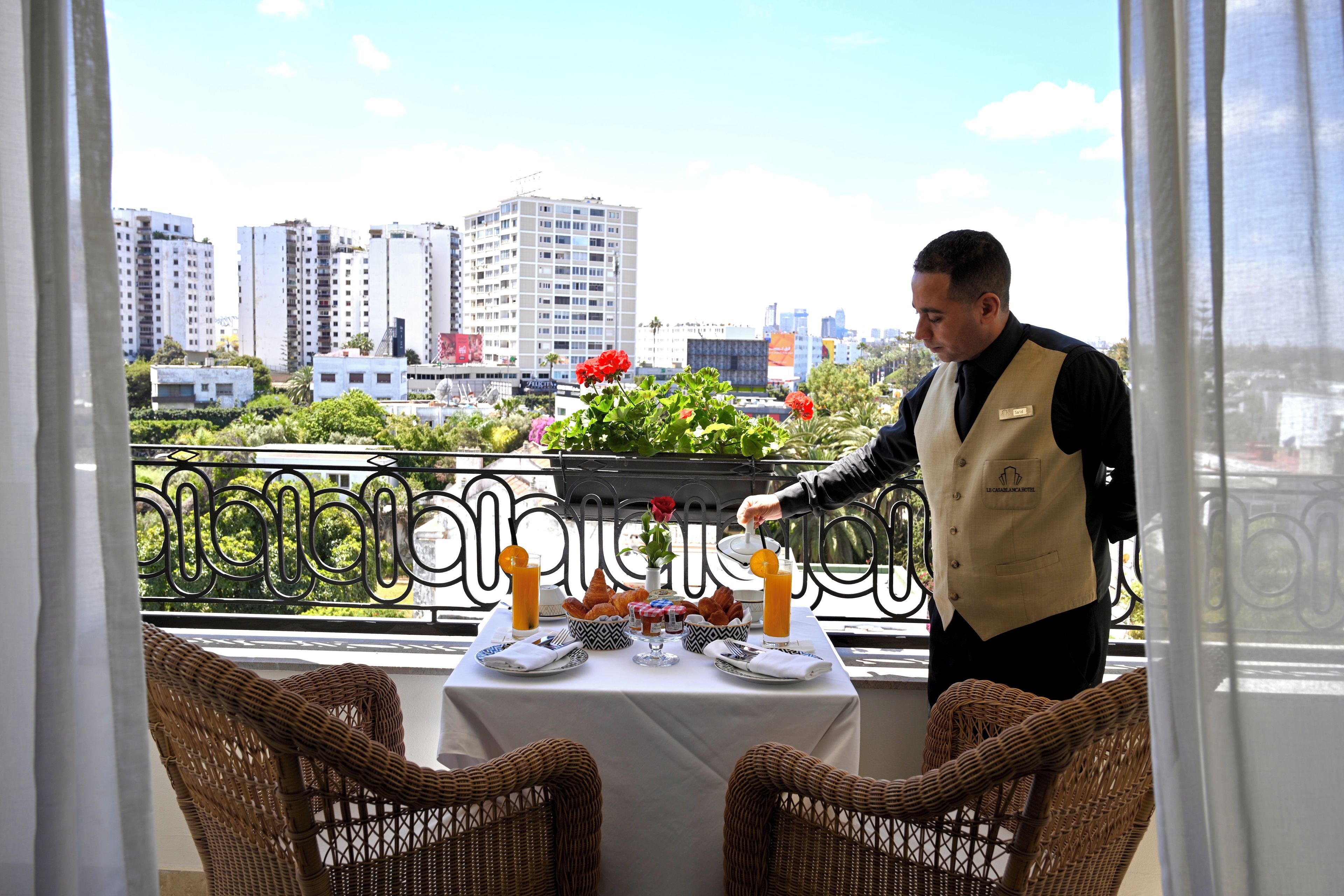 Une chambre d’hôtel avec une table sur le balcon, préparée pour le petit-déjeuner, avec des croissants, du jus d’orange et un vase rempli de fleurs.