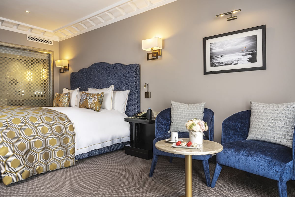 A hotel room with a large bed, a desk with a chair, a round table with a vase and a plate of strawberries, and a framed picture on the wall.