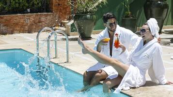 Una piscina con agua salpicando, dos personas con túnicas blancas, una sosteniendo una bebida, y un muro de ladrillo al fondo.