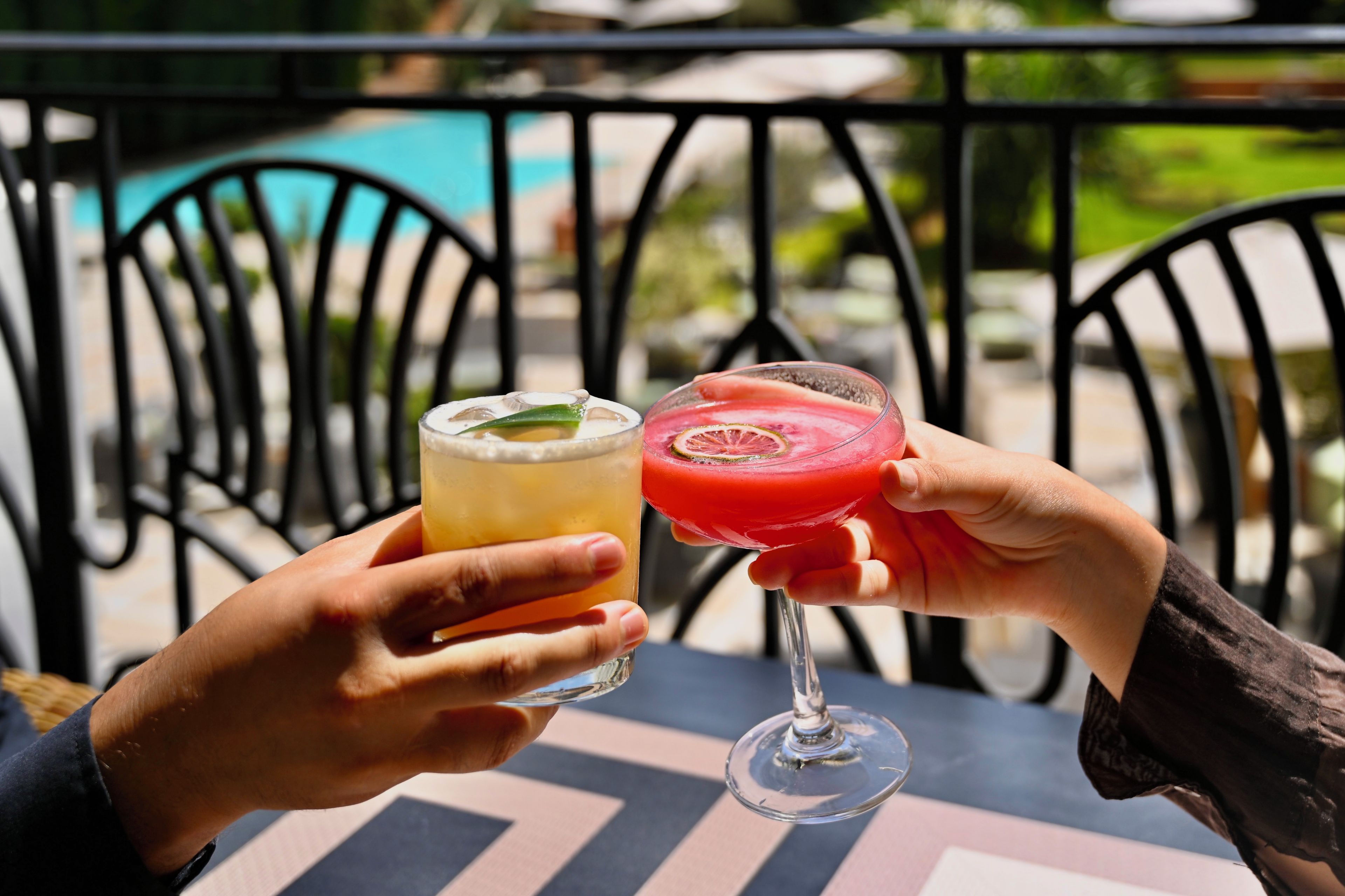 Two hands holding a glass of beer and a cocktail with a slice of fruit, outdoors with a pool and chairs in the background.