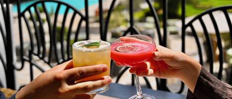 Two hands holding a glass of beer and a cocktail with a slice of fruit, outdoors with a pool and chairs in the background.