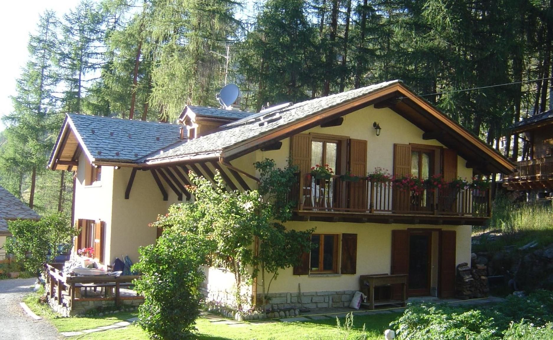 Une maison à deux étages avec un balcon, entourée de verdure et d’arbres.