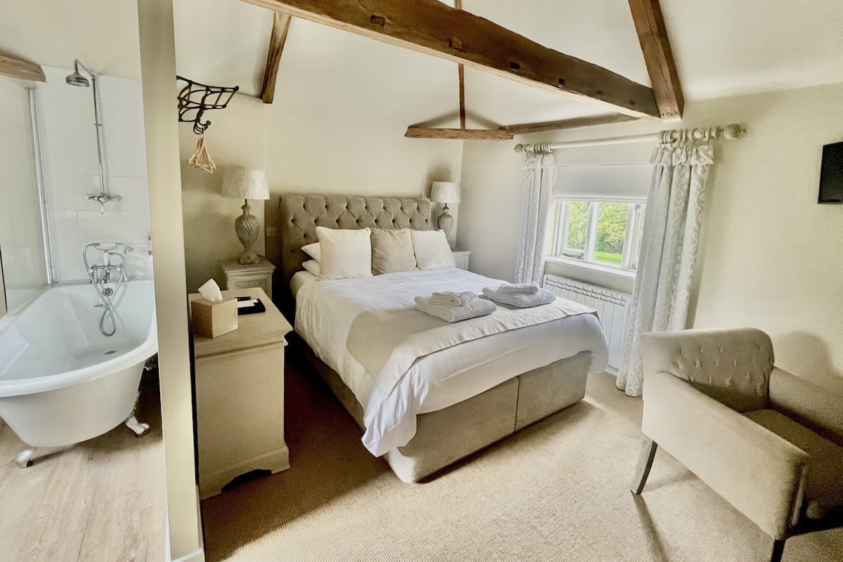 A bedroom with a large bed, a white bathtub, and a wooden ceiling structure.