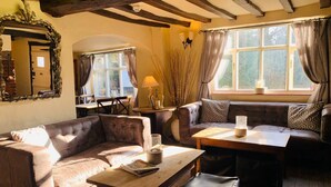 A cozy room with a brown leather sofa, a wooden table, and a bench. There is a window with curtains, a mirror, and a lamp on a side table. - The Kings Head Bawburgh (Norwich)