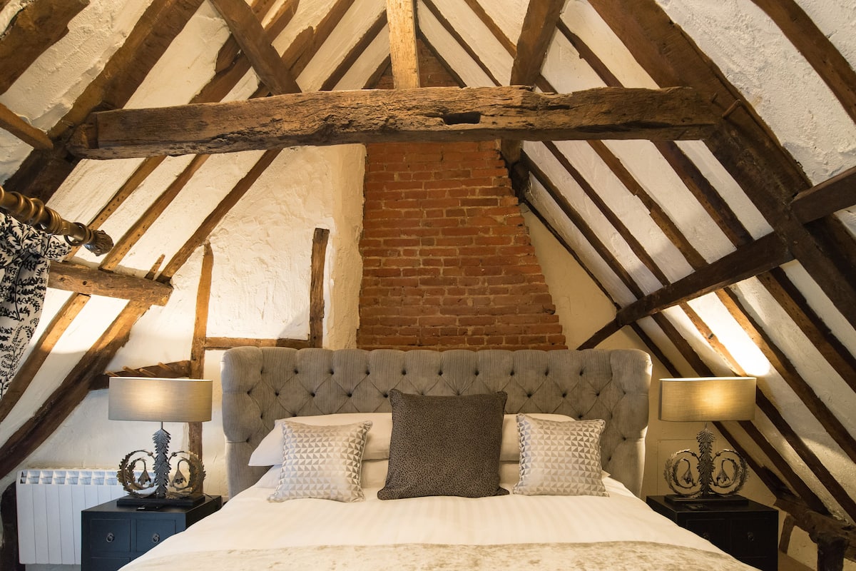A bedroom with a high ceiling, exposed wooden beams, a brick wall, a tufted headboard, and two bedside tables with lamps.