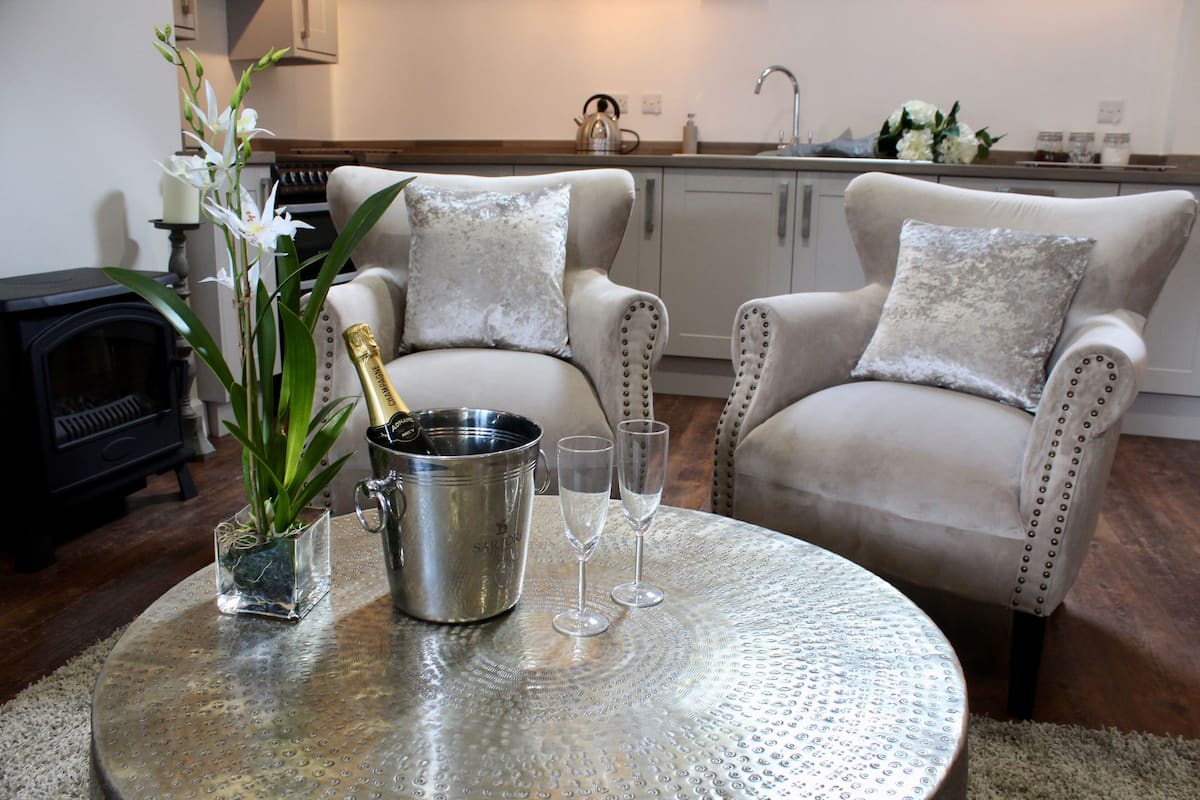 A living room with a round table, two armchairs, a fireplace, and a kitchen in the background.