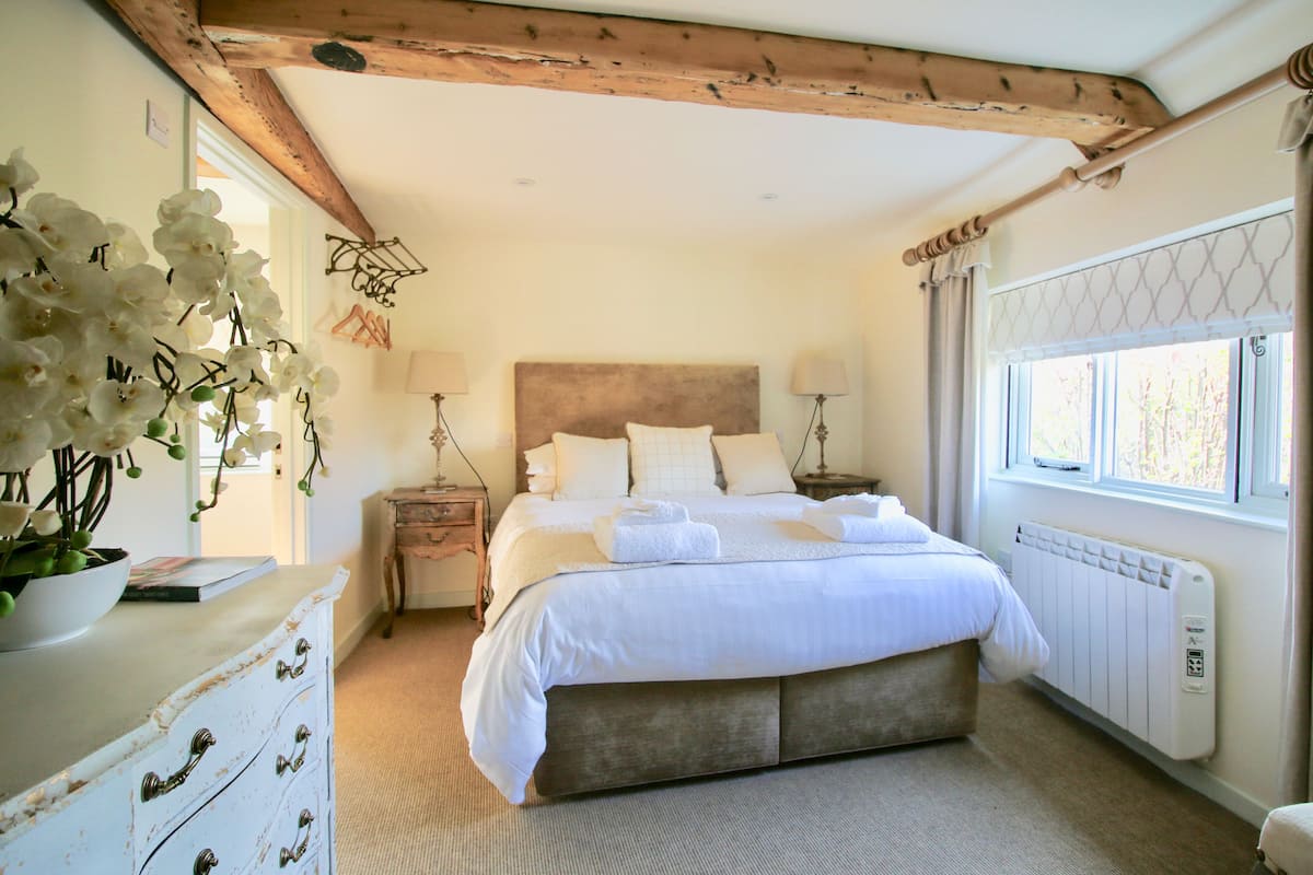 A bedroom with a large bed, wooden ceiling beams, a radiator, and a window with curtains.