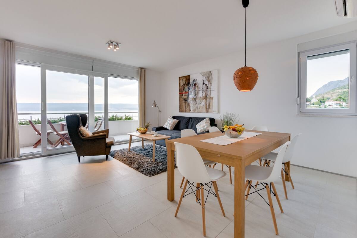 Ein modernes Wohnzimmer mit einer Couch, einem Esstisch und Stühlen. Ein großes Fenster bietet Blick auf das Meer und die Berge.