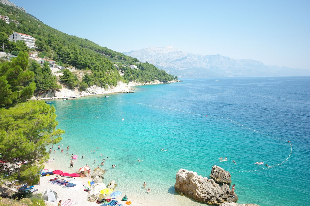 Ein Küstenstrand mit klarem türkisblauem Wasser, Felsformationen und einer Anhöhe mit Gebäuden.