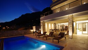 Un área de piscina moderna al aire libre con sillones de descanso e interiores bien iluminados.