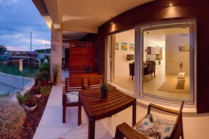 A modern outdoor patio with wooden furniture, a glass door leading to a kitchen, and a view of the surrounding area.