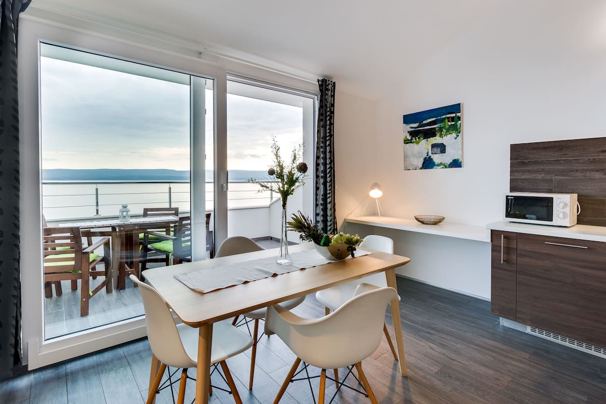 Ein modernes Zimmer mit Esstisch, Stühlen, einer Küchenzeile mit Mikrowelle und einem großen Fenster mit Meerblick.