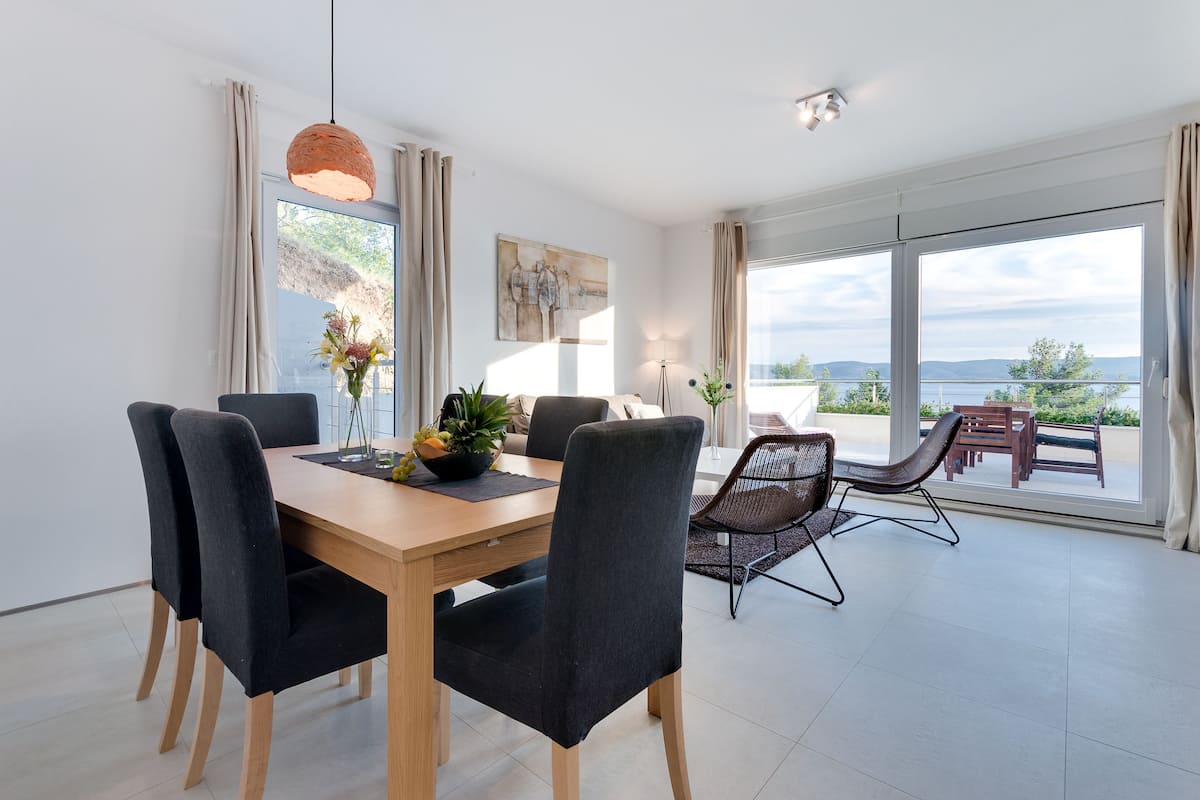 Ein stilvoller Essbereich mit einem Holztisch, schwarzen Polstermöbelstühlen und Blick ins Freie durch große Fenster.