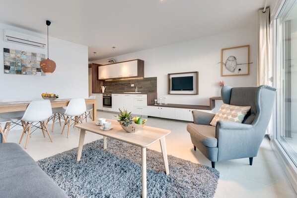 Una sala de estar moderna con sofá, mesa de centro y sillas. Cuenta con una zona de cocina con armarios y un televisor. Un ventanal grande proporciona luz natural.