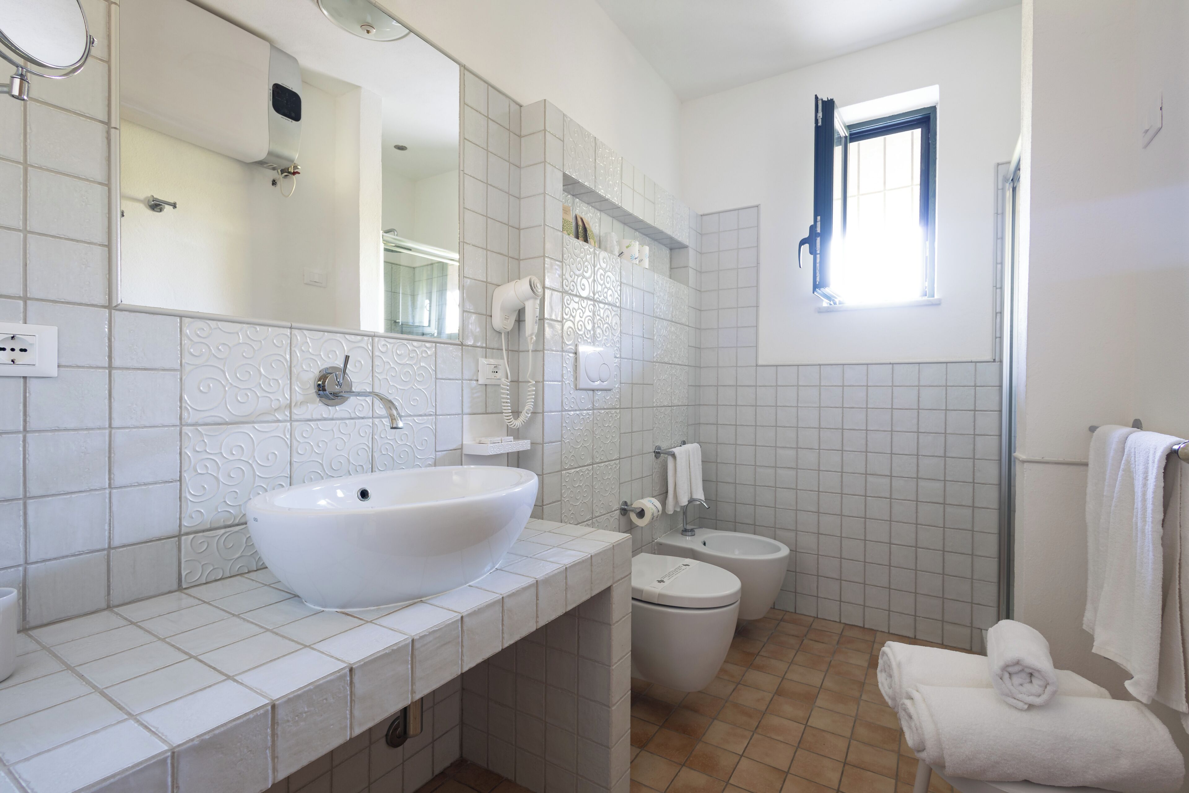 Ein Badezimmer mit einem großen, weißen Waschbecken, einem Spiegel, einer Toilette und einem Bidet. Ein Fenster bietet Blick ins Freie.