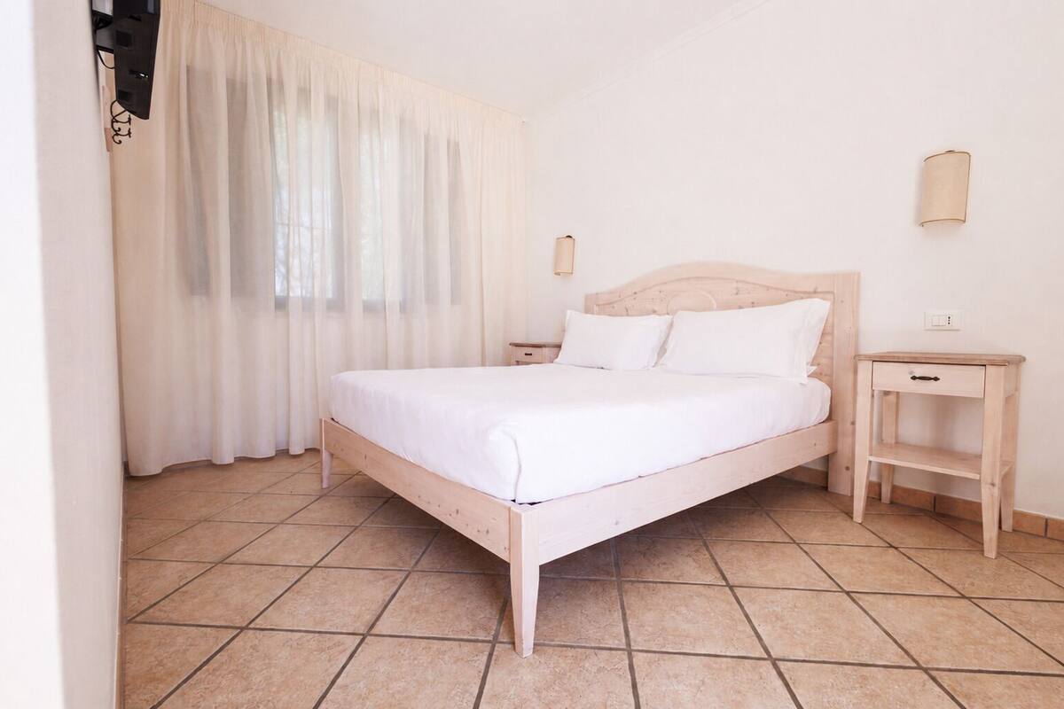 Un dormitorio con cama de madera, mesita de noche y cortinas translúcidas en la ventana.