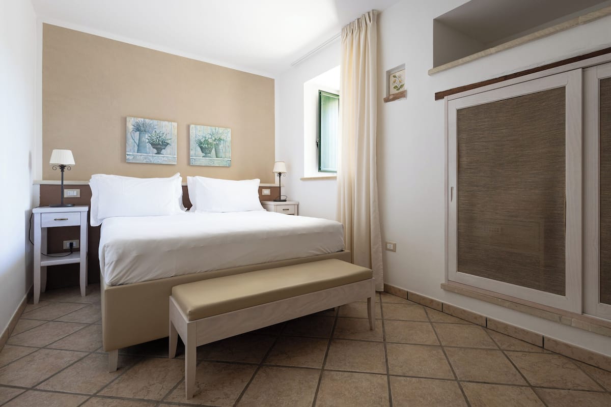Habitación de hotel con cama, mesitas de noche, un banco, una ventana con cortinas y una obra de arte en la pared.