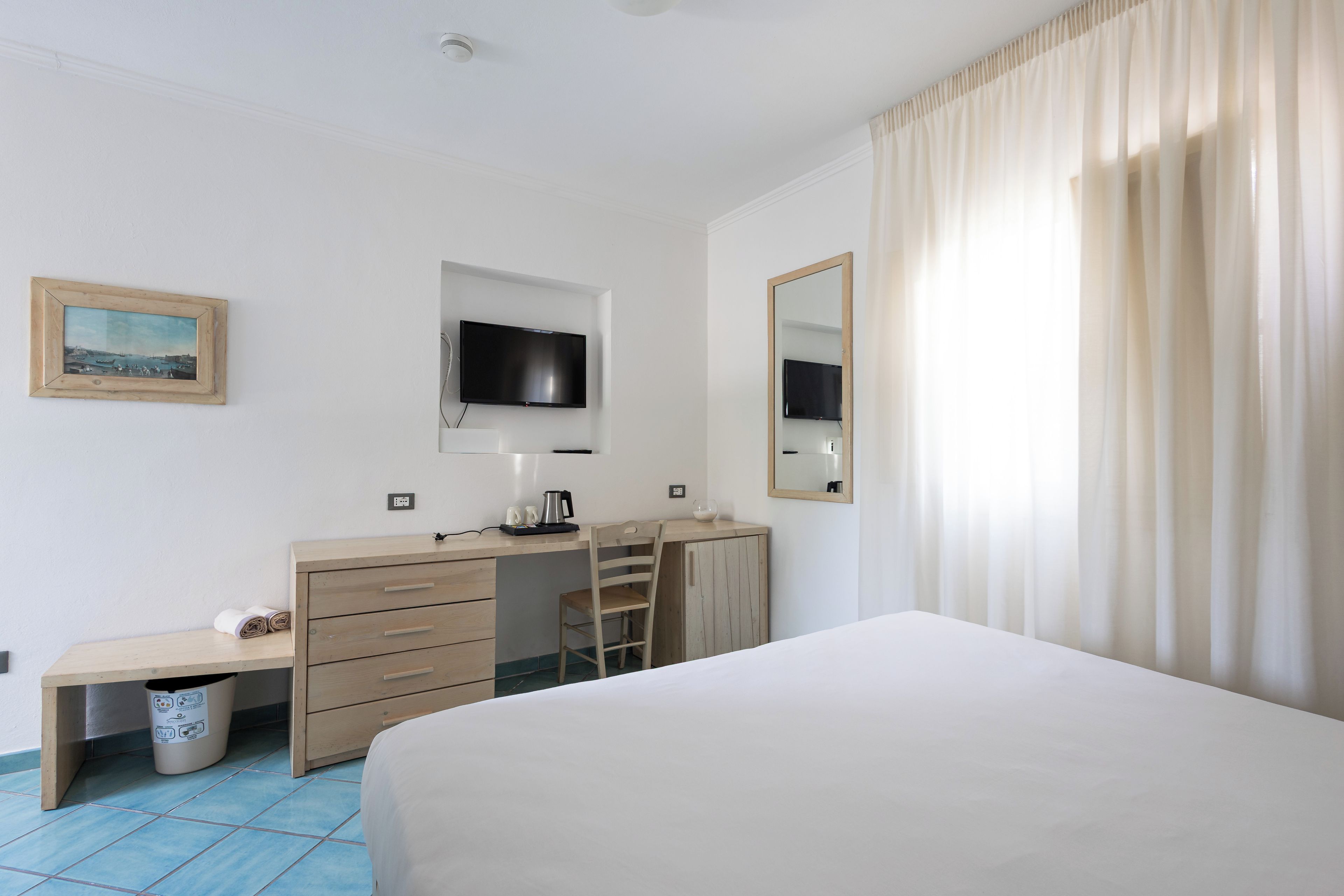 A hotel room with a bed, a desk with a chair, a television, a mirror, and a window with curtains.