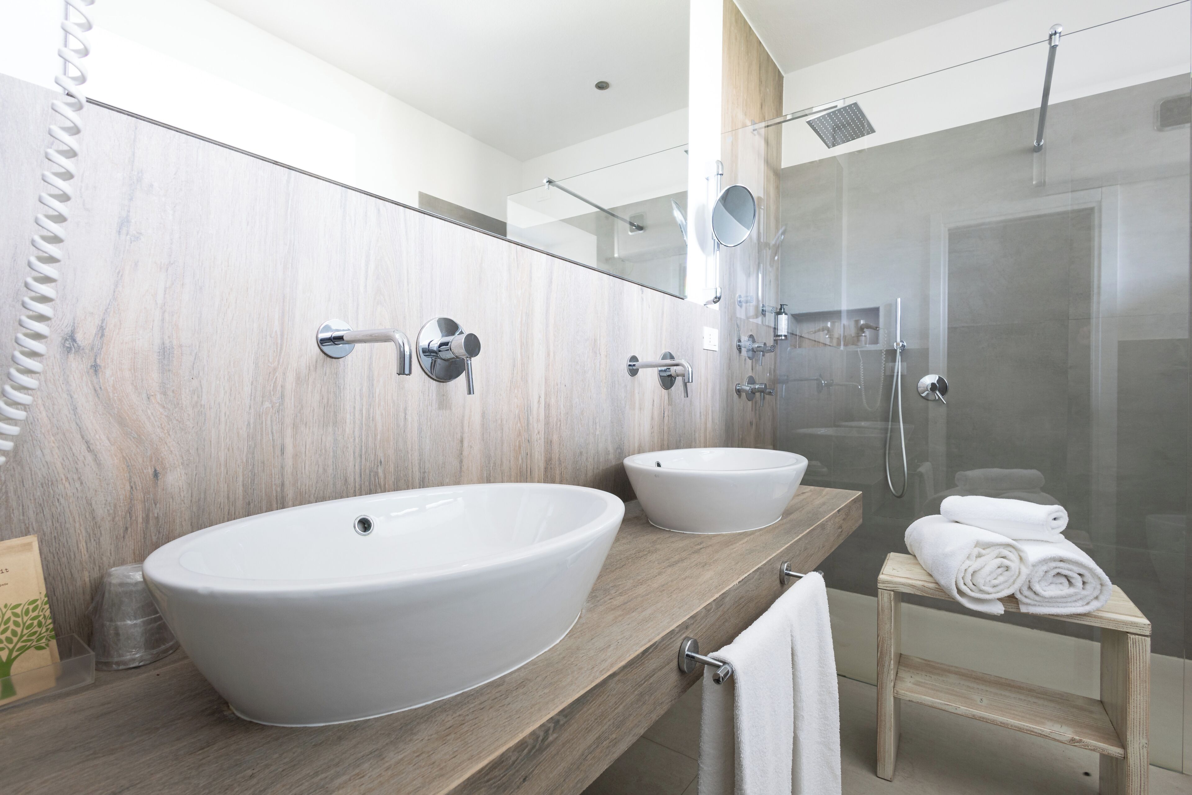 A modern bathroom with a large vessel sink, a smaller sink, a glass shower enclosure, and a wooden vanity.