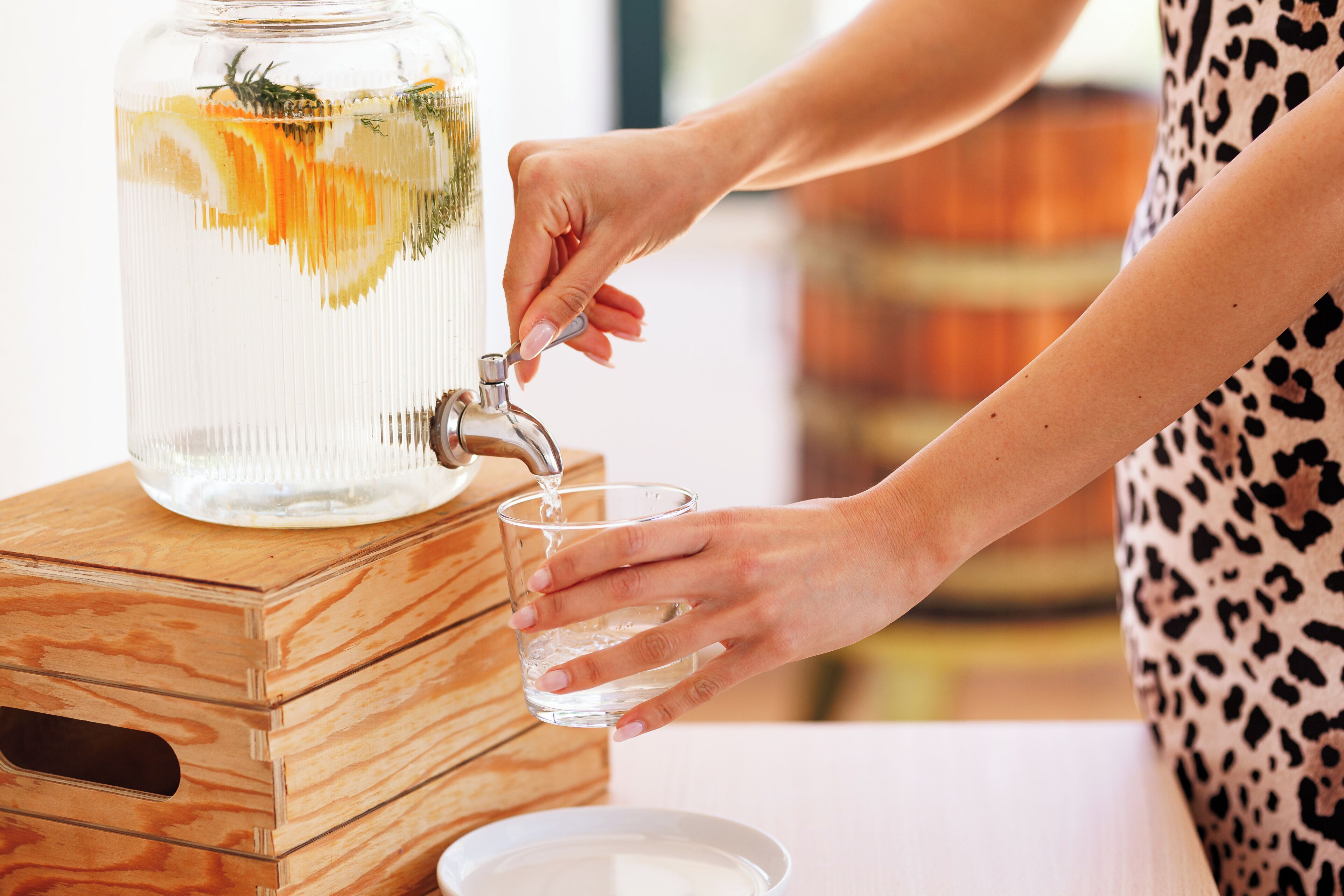A person filling a glass from a water dispenser with a clear container and herbs.