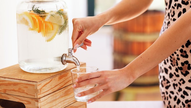 Eine Person füllt ein Glas an einem Wasserspender, der eine klare Kanne und Kräuter enthält.