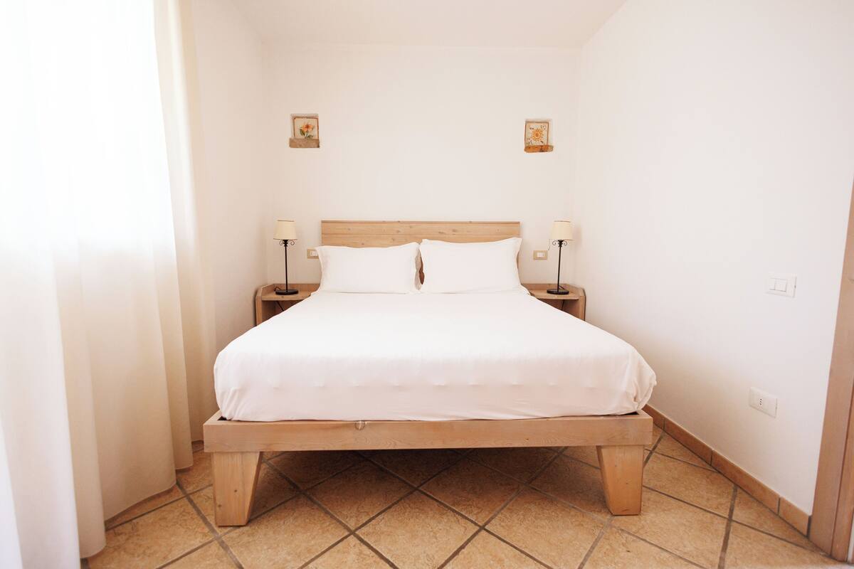 Una cama individual con estructura de madera y ropa de cama blanca en una habitación con paredes y suelo de baldosas blancos.