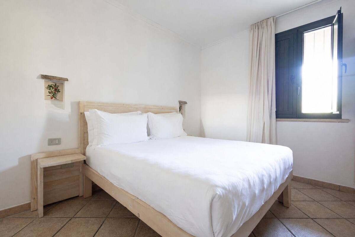 Un dormitorio con cama, cabecera de madera, mesita de noche, estante con una planta y ventana con cortinas, además de suelo de baldosas.