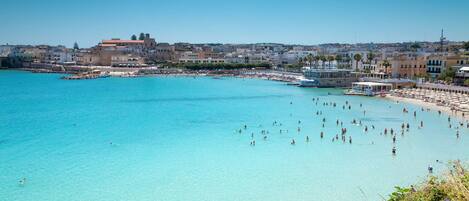 A coastal town with a clear blue sea, beach, and buildings along the shore.