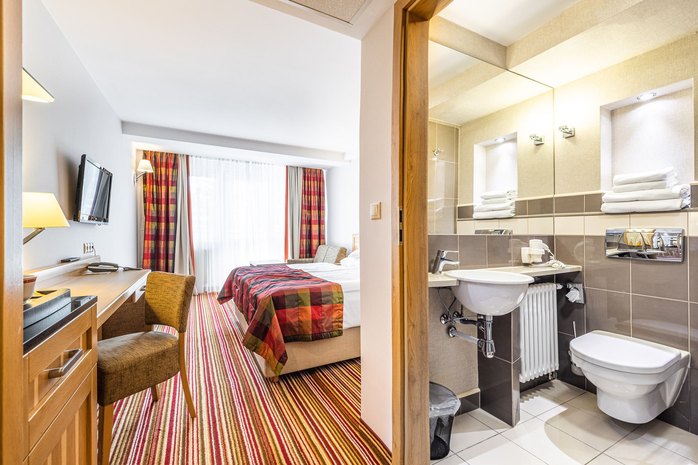 A hotel room with a bed, a desk with a chair, a TV, a window with curtains, and a bathroom with a sink, toilet, and mirror.
