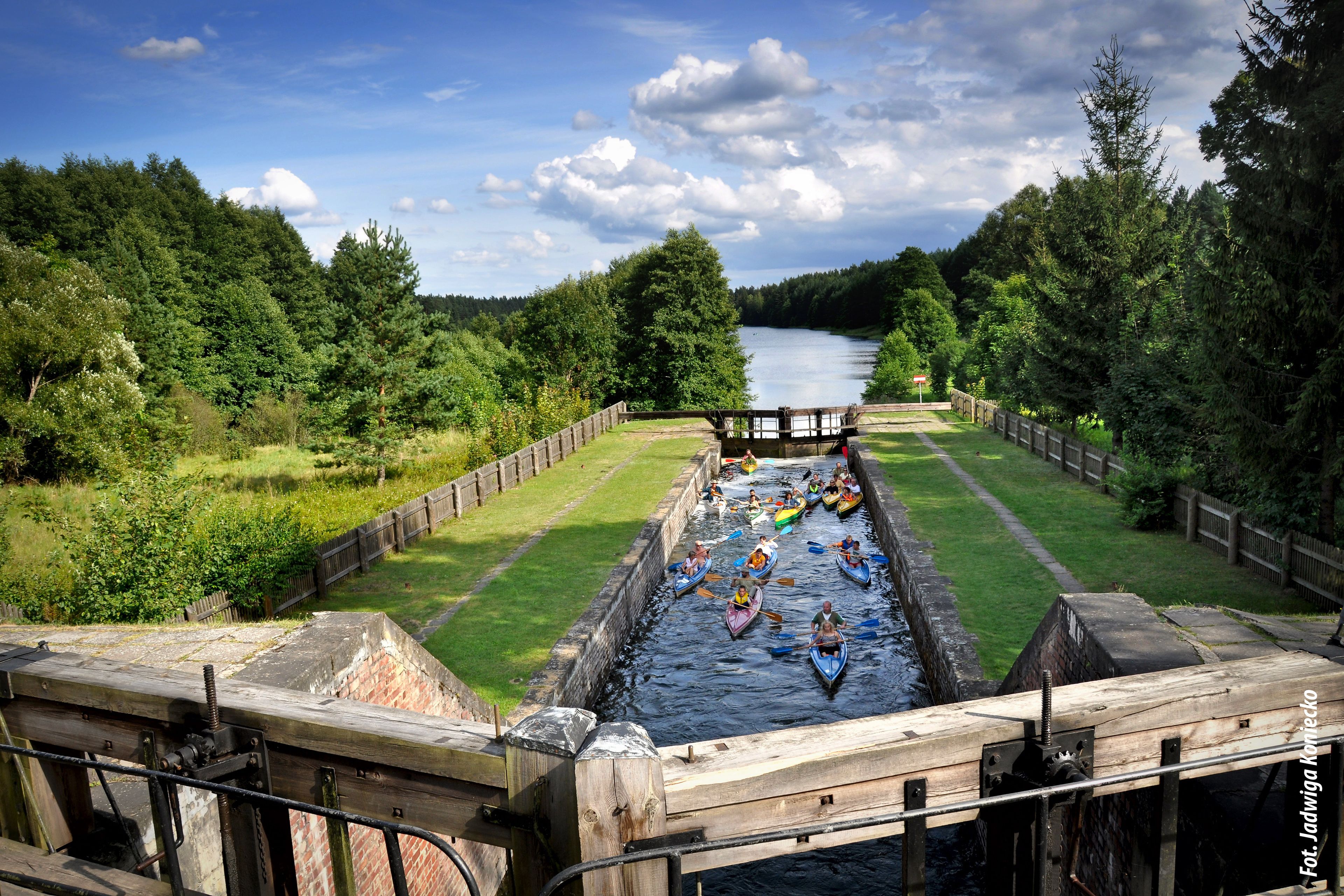 A river with canoes and kayaks, surrounded by greenery and a wooden bridge.