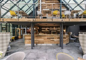 Zona bar moderna con elementi in legno, sedute con sedie gialle e grigie, e un soffitto in vetro.