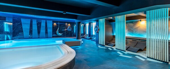 Un moderno espacio de spa con un amplio jacuzzi, zonas de descanso y una pared de madera con listones verticales.
