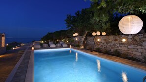 A swimming pool with stone walls and outdoor seating, surrounded by trees and stone lanterns. - Amalgam Homes Paros Beachfront Villa (Paros)
