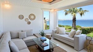 A modern living room with a large window overlooking the sea, a sofa, a coffee table, and decorative wall art. - Amalgam Homes Paros Beachfront Villa (Paros)