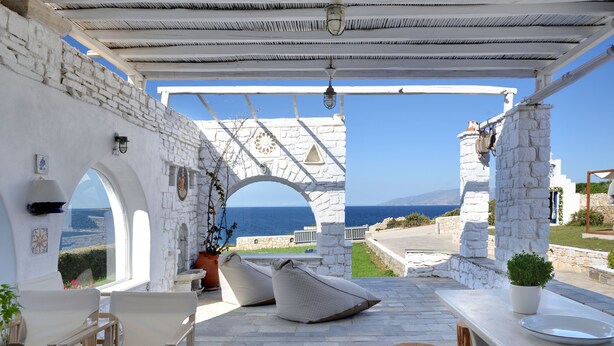 A covered outdoor seating area with stone walls, a view of the sea, and a stone archway. - Amalgam Homes Paros Beachfront Villa (Paros)
