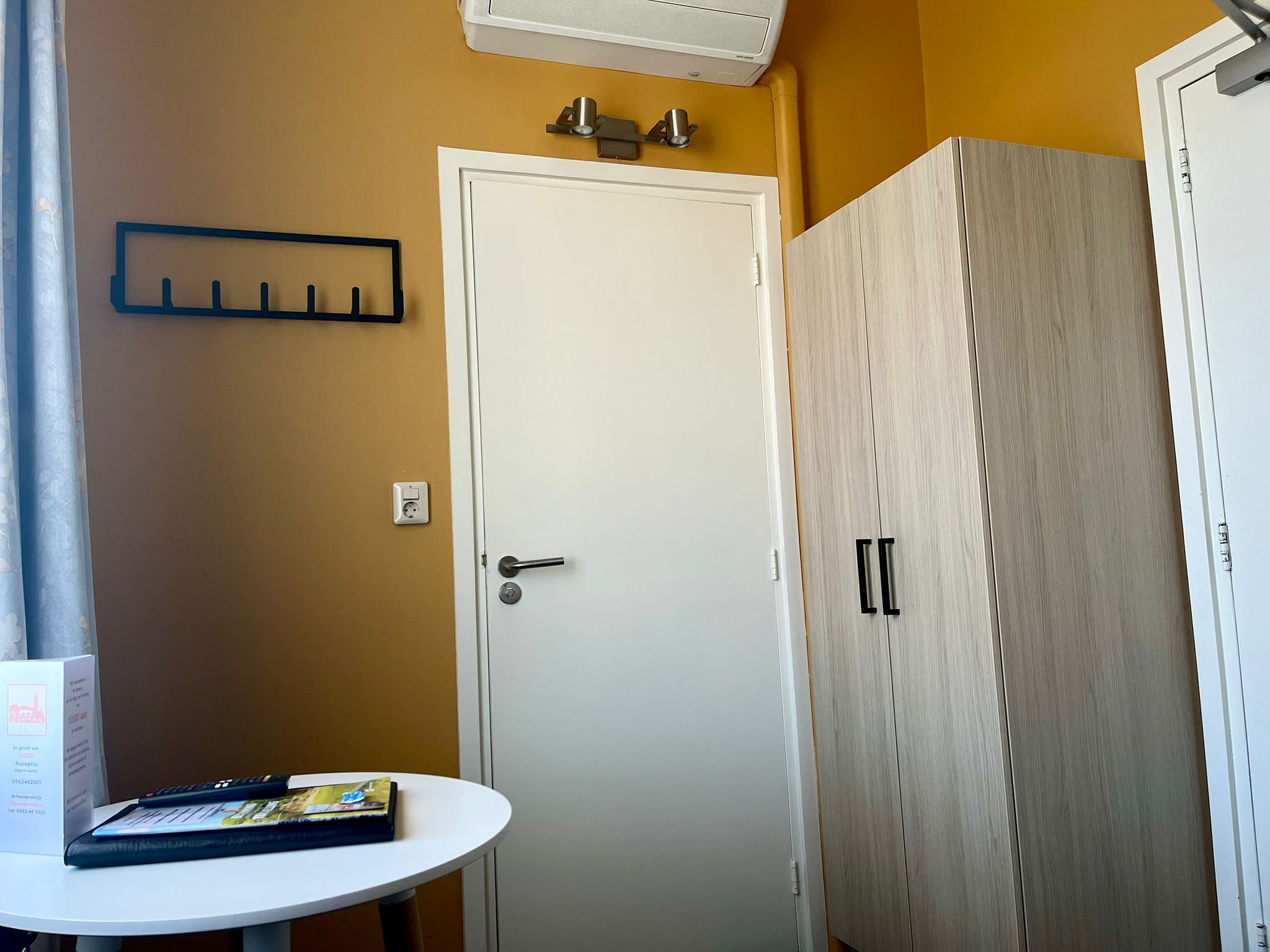 Ein Zimmer mit weißer Tür, einem Holzschrank, einem kleinen Tisch mit einem Buch und einem an der Wand befestigten Lichtschalter.